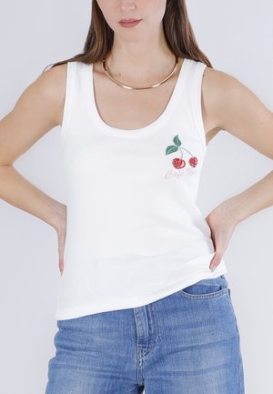 Femme portant un haut sans manches blanc à côtes avec des cerises rouges brodées et des feuilles vertes, assorti à un jean bleu et un collier doré.