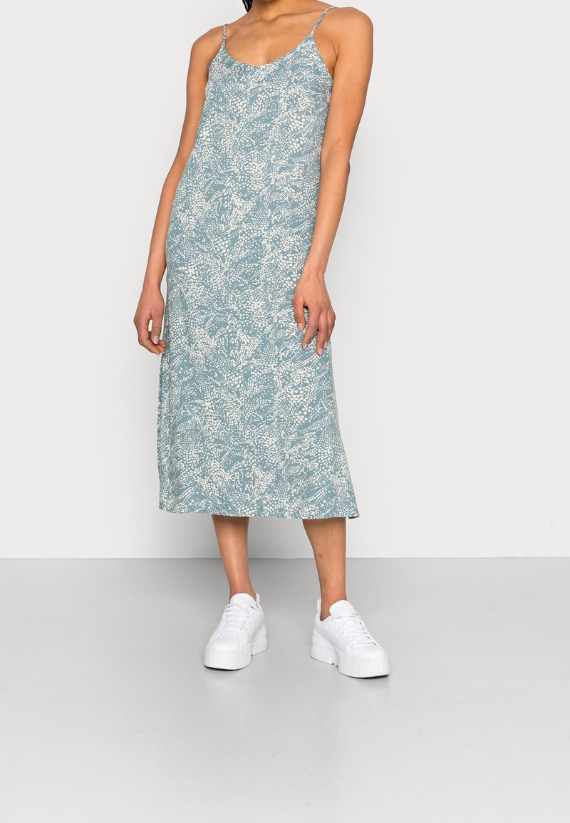 Mujer con un vestido midi de patrón abstracto en azul claro y blanco, con tirantes finos y zapatillas de plataforma blancas, de pie frente a un fondo liso.