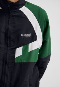 Črna in zelena športna jakna z belimi poudarki. Ima visok ovratnik, zadrgo ter logo "hummel sportswear" na prsih. Gladka textura.