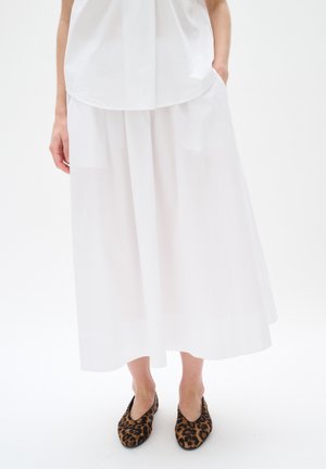Person wearing a white midi skirt and white top paired with leopard print flat shoes standing with one hand in pocket.