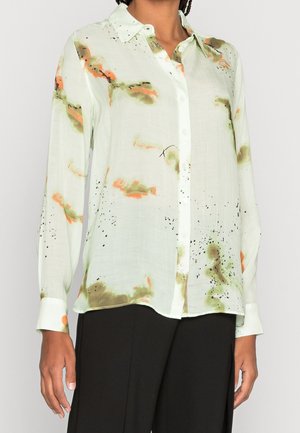 Femme portant un chemisier à manches longues, boutonné, de couleur claire avec des motifs abstraits orange et verts, associé à un pantalon noir.