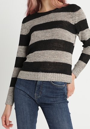 Femme portant un pull en tricot rayé horizontal noir et gris et un jean en denim bleu, debout avec une main dans la poche.