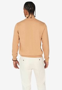 Tan brei-sweater met een hoge kraag en geribde manchetten, gedragen met witte broeken. De stof lijkt glad en sluit nauw aan op het lichaam.