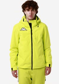 Giacca neon gialla brillante con chiusura a zip frontale, cappuccio regolabile, polsini elasticizzati e logo Kappa sul petto. Materiale liscio e leggero.