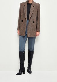 Nainen, jolla on päällään ruskea herringbone-takki, musta paita, siniset housut ja mustat polvipituiset korkokengät, seisoo tasaisella taustalla.