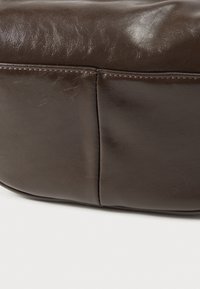 Geantă din piele maro închis cu textură netedă, formă rotunjită și cusături vizibile. Prezintă un detaliu de cusătură de-a lungul părții.