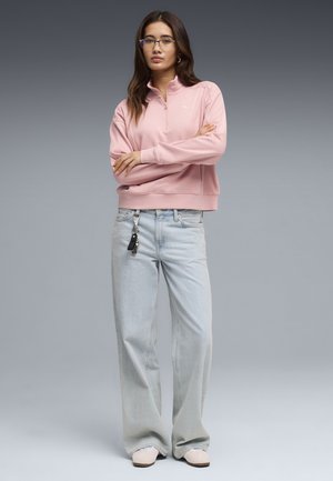 Jonge vrouw met lang haar en een bril staat met gekruiste armen, gekleed in een roze sweatvest met rits, lichtblauwe jeans met wijde pijpen en beige schoenen.