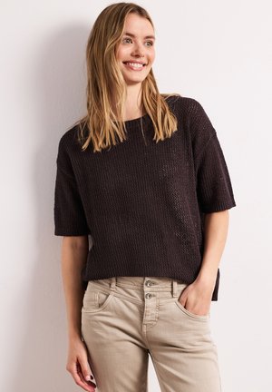 Smilende kvinde med blondt hår iført en sort tekstureret kortærmet sweater og beige bukser, den ene hånd i lommen, mod en ensfarvet baggrund.