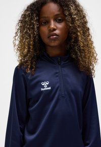 Mörkblå atletisk zip-up-top i mjukt, lätt tyg med hög krage och vit logotyp på vänster bröst.