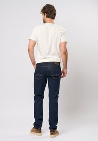 Jeans oscuras de corte recto, combinados con una camiseta de color claro. Incluye bolsillos traseros y una etiqueta en la cintura. Sin patrones visibles.