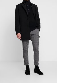 Czarny płaszcz na granatowym swetrze z dzianiny, szare spodnie w kratę i czarne zamszowe botki. Płaszcz ma stojący kołnierz i zapięcie na guziki.