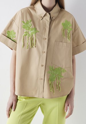 Beige kortærmet skjorte med knapper og grøn blomster palietbroderi, båret med lysegrønne bukser på en stående person.