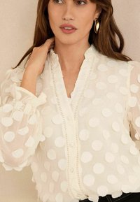 Blusa trasparente color crema con pois bianchi a trama, colletto arricciato e chiusura frontale con bottoni, dotata di maniche con polsini e dettagli in pizzo.