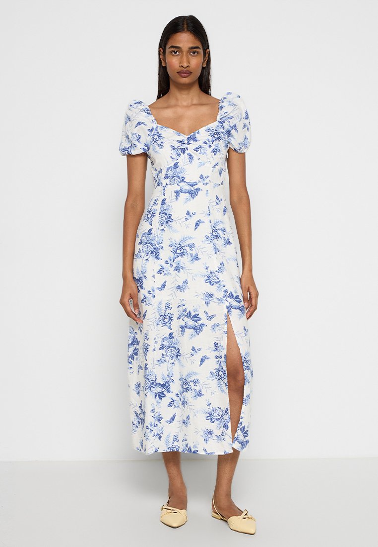 Weißes Blumenkleid mit blauen botanischen Mustern, kurzen Puffärmeln, einem quadratischen Ausschnitt und einem seitlichen Schlitz für zusätzliche Details.