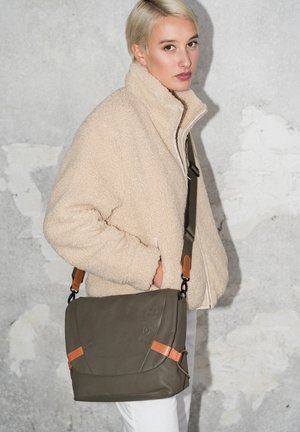 Borsa a tracolla verde oliva in pelle testurizzata con dettagli color cuoio e tracolla regolabile. Il modello indossa una giacca in pile crema, in piedi contro un muro grigio.