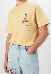 Yellow cotton t-shirt featuring a black print of a cockatoo with flames. Paired with distressed light blue denim shorts.