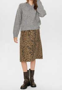 Žena v šedém pleteném svetru, midi sukni s leopardím potiskem a tmavě hnědých kožených botách sahajících po lýtka, stojící proti jednobarevnému pozadí.