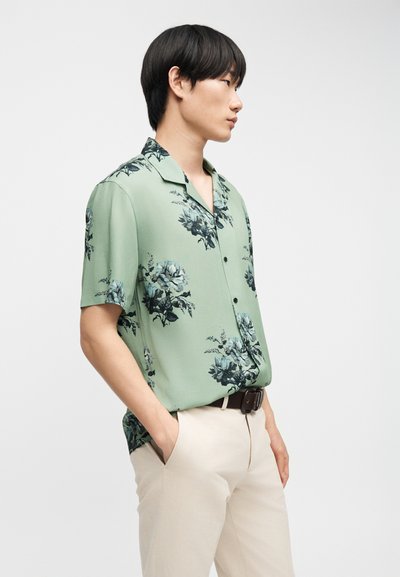 Νεαρός άνδρας με μαύρα μαλλιά που φοράει ένα ανοιχτό πράσινο φλοράλ πουκάμισο με κοντά μανίκια, φουστανωμένο σε μπεζ παντελόνι, με τα χέρια στις τσέπες.