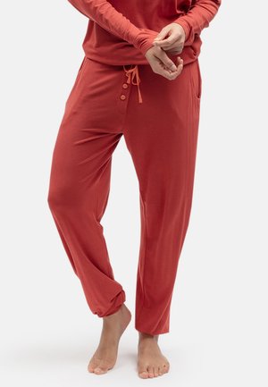 Persoon die losse rode loungebroek draagt met knoopdetail en trekkoord, staat blootsvoets en past de mouw van een shirt met lange mouwen aan tegen een effen achtergrond.