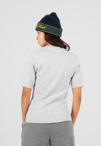 Donna di spalle che indossa una maglietta grigia a maniche corte, pantaloni grigi e un berretto blu navy e verde con testo giallo.