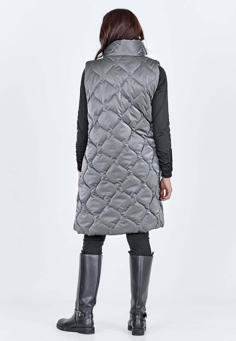 Vrouw die een lange grijze doorgestikte mouwloze jas draagt, een zwart shirt met lange mouwen, een zwarte broek en zwarte kniehoge laarzen, staand met haar rug naar voren.