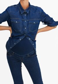 Camisa de mezclilla en azul oscuro con cierre de botones en la parte delantera, cuello y dos bolsillos en el pecho. Combinada con pantalones vaqueros oscuros a juego, creando un look cohesivo.