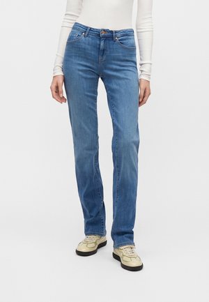 Person, die hellblaue Jeans mit geradem Bein, ein weißes Langarmshirt und beigen Sneakers mit schwarzen Sohlen trägt, steht vor weißem Hintergrund.