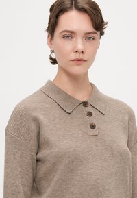 Beige ribbed knit polo with a collar, three dark brown buttons, and a relaxed fit. Fabric shows subtle texture and warmth.