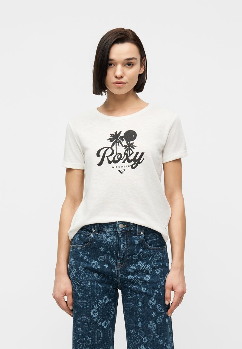 Νεαρή γυναίκα με κοντά σκούρα μαλλιά, φοράει άσπρο μπλουζάκι με γραφικό σχέδιο Roxy και σκούρο μπλε τζιν με λουλουδάτο μοτίβο, κοιτάζει μπροστά.