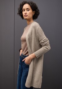 Beige dlouhý kardigan vyrobený z pleteného materiálu, s ribbenovými manžetami a otevřeným předním dílem. Nositelný přes hnědý top a modré džíny.