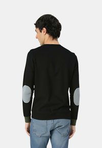 Maglione nero con maniche lunghe, scollatura rotonda, toppe grigie sui gomiti e polsini a coste verdi. Il materiale sembra liscio ed elastico.