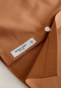 Pruun puuvillane särk, millel on nööbid ja õmmeldud silt, millel on kiri "DURABLE GOODS LOT 082." Sile tekstuur, kortsunud krae serv on nähtav.