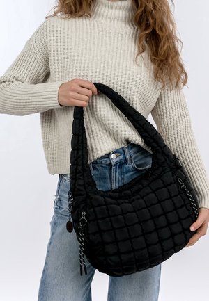 Frau trägt cremefarbenen Strickpullover und blaue Jeans, hält große schwarze gesteppte Handtasche mit gepolstertem Griff vor weißem Hintergrund.