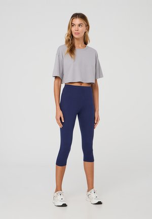 T-shirt grigio corto con maniche corte abbinato a leggings capri blu scuro. Il modello indossa sneakers bianche, dal design semplice e dalla texture liscia.