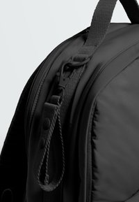 Close-up of a black backpack strap with a plastic clip and two black snap buttons on a fabric loop near a zipper.