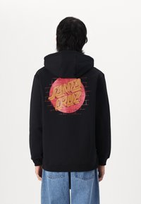 Črna mikica s kapuco, ki na hrbtni strani prikazuje živahno rdečo in rumeno grafiko "Santa Cruz" na vzorcu zidane stene.