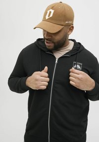 Crna zip-up dukserica s prednjim džepom i logotipom, nosi se preko bež majice. Osoba nosi smešnu kapu s belim logotipom.