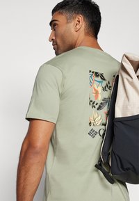 Lys grønn bomullst-skjorte med korte ermer, med et livlig blomstermotiv på ryggen. Mannen holder en beige og svart ryggsekk.