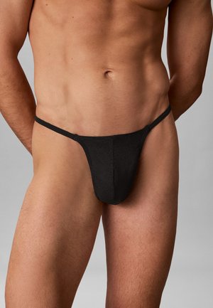 Männlicher Torso trägt schwarze minimal bedeckende Slip-Unterwäsche mit schlanken seitlichen Trägern vor einfachem Hintergrund.