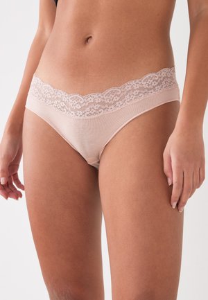 Nainen pukeutune vaaleanpunaiseen pitsireunaiseen alusvaatteeseen, paljastaen vatsan ja reisien yläosan vaaleaa yksinkertaista taustaa vasten.