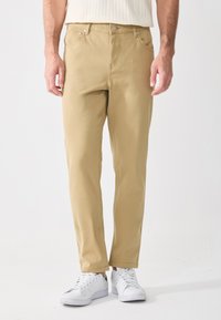 Beige puuvillahousut suoran muotoilun kanssa, viisi taskua ja kontrastiväriset tikkaus, yhdistettyinä valkoisiin tennareihin.