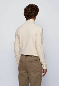 Man with curly brown hair wearing a cream long-sleeve shirt and brown pants, standing with his back facing the camera against a plain backdrop.