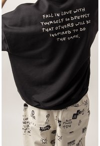 Czarna bawełniana koszulka z białym nadrukiem na plecach. Połączona z kremowymi spodniami z czarnymi grafikami w stylu doodle i różnymi motywami.