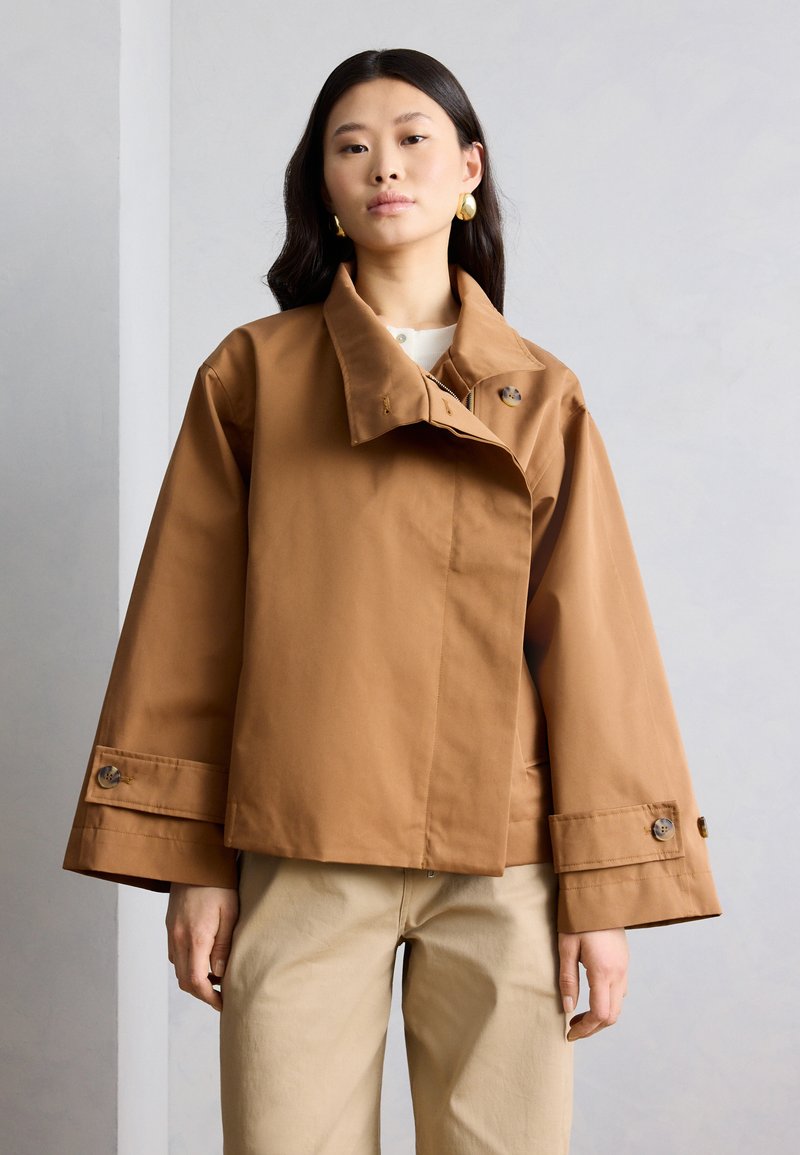 Femme portant une veste marron oversized à manches larges et un pantalon beige, debout devant un fond uni gris clair.