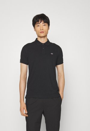 Homme portant un polo noir à manches courtes avec un petit logo brodé sur la poitrine gauche, debout devant un fond blanc uni.
