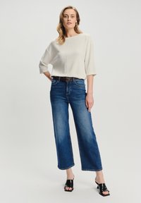 Donna in piedi che indossa un top a coste color crema con maniche a 3/4, jeans blu a vita alta e mules con tacco aperti neri su sfondo bianco.