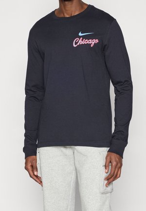 Homme portant une chemise noire à manches longues avec un logo Nike bleu et un texte rouge "Chicago", associé à un pantalon cargo gris clair.