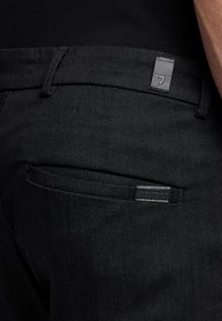 Jeans in denim nero con una superficie liscia. Le caratteristiche includono un'etichetta con la scritta '7' e due tasche posteriori dal design minimalista.