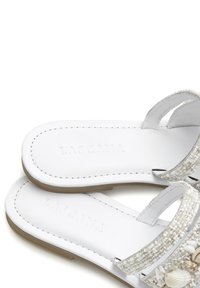 White sandals with two narrow straps featuring bead and shell embellishments. Smooth leather-like texture, flat sole, and embossed logo.