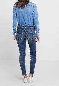 Jeans skinny in denim blu scuro con vita alta, tasche posteriori e orli delle caviglie sfilacciati, abbinati a una camicia in denim blu chiaro.